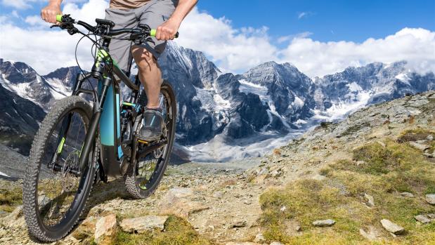 Ein Mountainbiker fährt mit einem Fahrrad auf einem steinigen Bergpfad vor verschneiten Alpen.