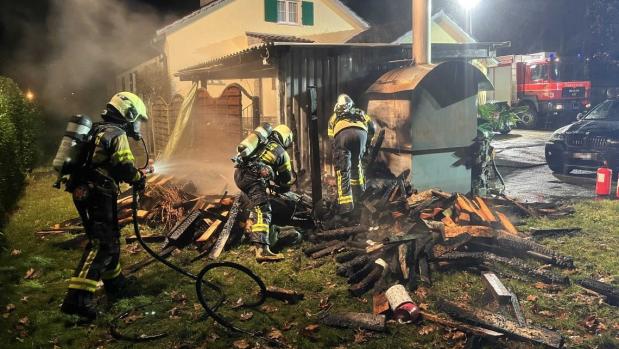 Feuerwehrleute löschen einen Brand an einem Holzstapel neben einem Haus bei Nacht