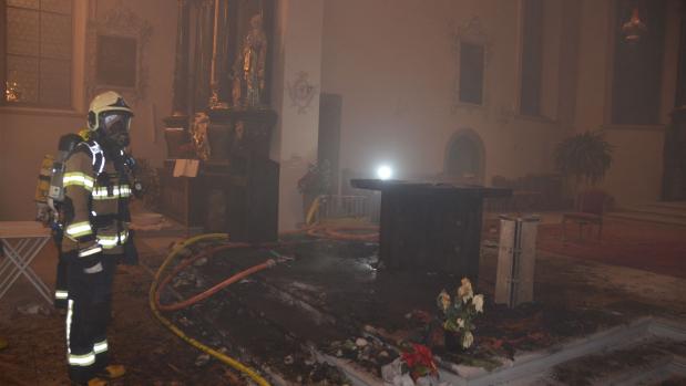 Ein Feuerwehrmann in Schutzausrüstung steht in einer verrauchten Kirche neben verbrannten Trümmern und Schläuchen.