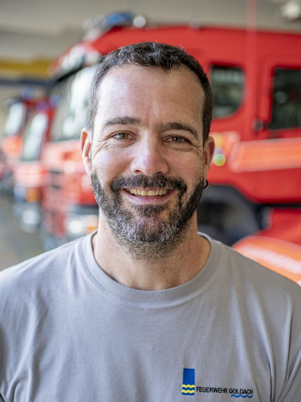 Lächelnder Mann in einem grauen T-Shirt vor roten Feuerwehrfahrzeugen in einer Halle