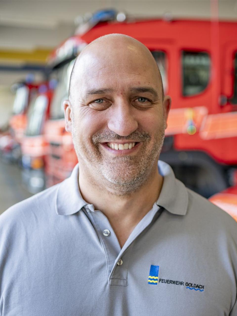 Lächelnder Mann in grauem Polohemd vor roten Feuerwehrfahrzeugen in einer Halle