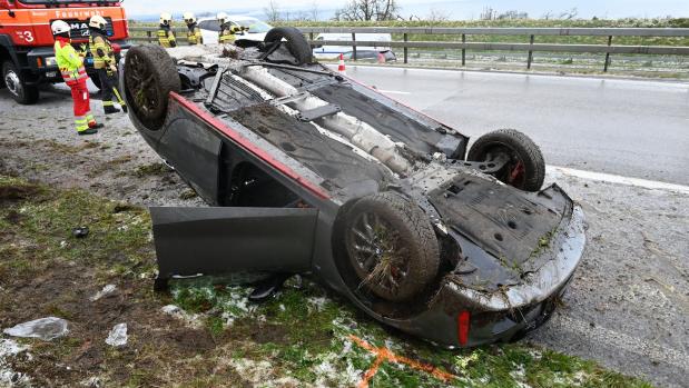 Umgekipptes Auto nach einem Unfall am Straßenrand, daneben stehen Feuerwehrkräfte und ein Löschfahrzeug.